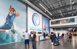 Visitors and athletes inside the Virginia Beach Sports Center, featuring a large mural of a basketball player, modern design elements, and the venue's logo.